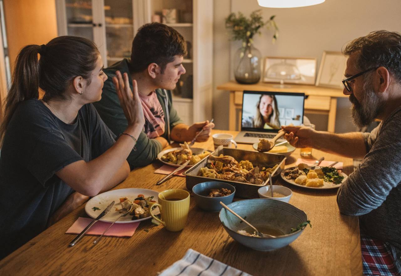 Family sitting at dinner table eating and interacting with a female on a video call shown on laptop